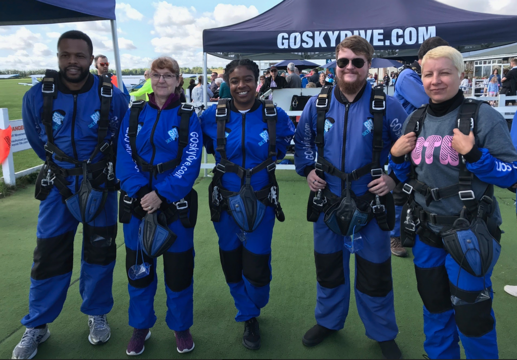 A group of five people stand in skydiving harnesses, smiling at the camera.